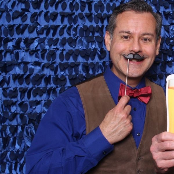 Man in photo booth holding up fake black mustache to face